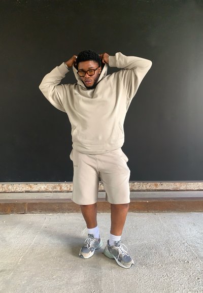 Conjunto de sudadera beige y pantalones cortos, con un acabado texturizado. Zapatillas grises con acentos. Poses individuales con las manos en la sudadera. Fondo liso.