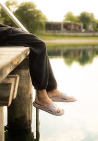Svetlo šedé sandále typu slide otvorenou špičkou s ventilačnými otvormi a textúrovanou podrážkou, nosené s čiernymi joggingovými nohavicami, odpočívajúce nad vodou na móle.
