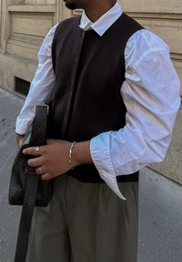 Tumši brūns žakets virs baltā pogājamā krekla, kopā ar brīviem bēšiem biksēm. Strukturēts melns soma turēts vienā rokā, sudraba gredzeni akcentē pirkstus.