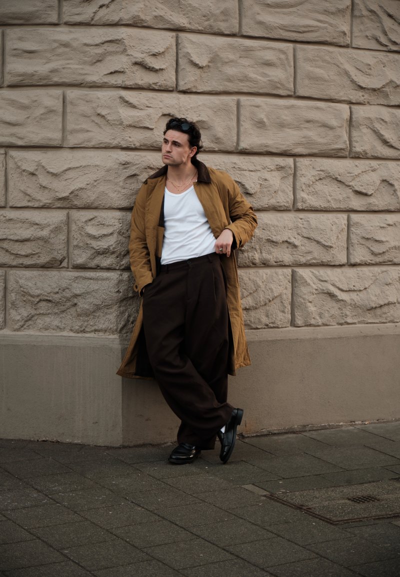 Homme en long manteau marron, chemise blanche et pantalon ample foncé appuyé contre un mur en pierre texturé sur un trottoir en ville.