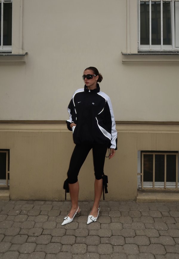 Frau trägt eine schwarz-weiße Sportjacke, schwarze kurze Leggings mit Schleifen, weiße spitze Schuhe und eine Sonnenbrille und steht auf einem Kopfsteinpflaster.