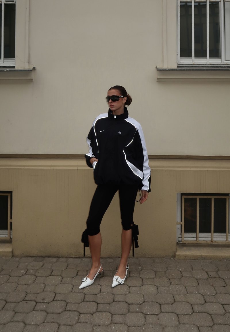 Femme portant une veste de sport noire et blanche, des leggings courts noirs avec des nœuds, des chaussures blanches pointues et des lunettes de soleil, debout sur un trottoir en pavés.