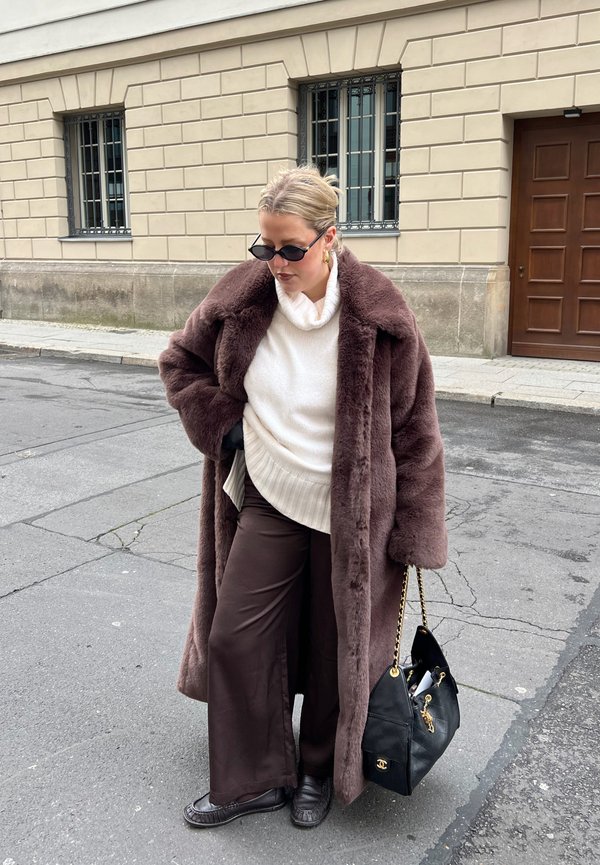 Mujer con abrigo de piel marrón oscuro, suéter blanco, pantalones marrón oscuro, zapatos negros, gafas de sol, llevando un bolso negro en una calle de la ciudad.