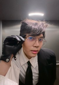 Jeune personne portant des lunettes et un gant en cuir noir ajuste ses lunettes, vêtue d'une chemise blanche, d'une cravate noire et d'une veste dans un ascenseur métallique.