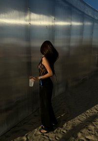 Μαύρη δαντελωτή μπλούζα, μαύρο παντελόνι φάρδειας, και σανδάλια. Κρατώντας ένα διάφανο ποτήρι, στέκοντας σε άμμο κοντά σε μια ανακλαστική, κυματοειδή τοιχοποιία.