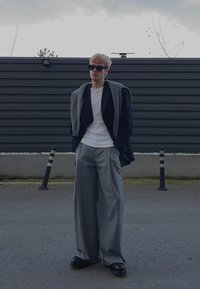 Jeune personne portant des lunettes de soleil noires, chemise blanche, blazer noir avec un pull gris drapé sur les épaules, pantalons larges gris, debout à l'extérieur.