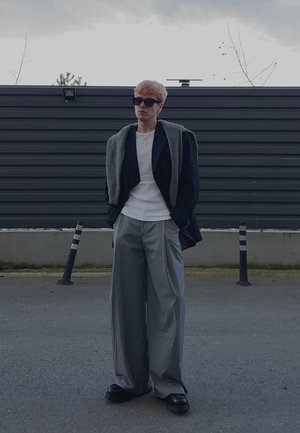Joven con gafas de sol negras, camisa blanca, blazer negra con suéter gris sobre los hombros, pantalones grises anchos, de pie al aire libre.