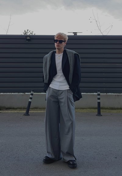 Joven con gafas de sol negras, camisa blanca, blazer negra con suéter gris sobre los hombros, pantalones grises anchos, de pie al aire libre.