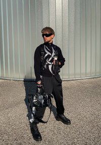 Črna strukturalna zip-up jopica s srebrnim vzorcem, črne hlače, debeli črni škornji, rokavice brez prstov in sijoča črna torba.