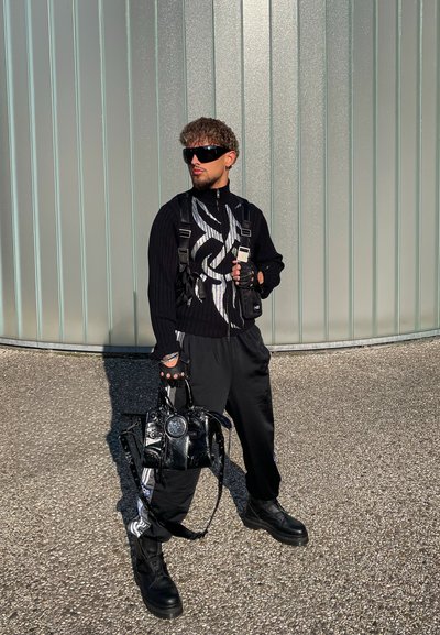 Suéter negro de canalé con cremallera y patrón plateado, pantalones negros, botas negras chunky, guantes sin dedos y un bolso negro brillante.