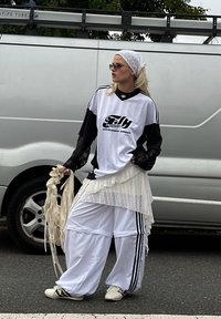 Oversized svart-hvitt sportskjorte med logo, lagdelt hvit skjørt, løse hvite bukser med svarte striper, og hvite sko. Tilbehør inkluderer et hodebånd og solbriller.
