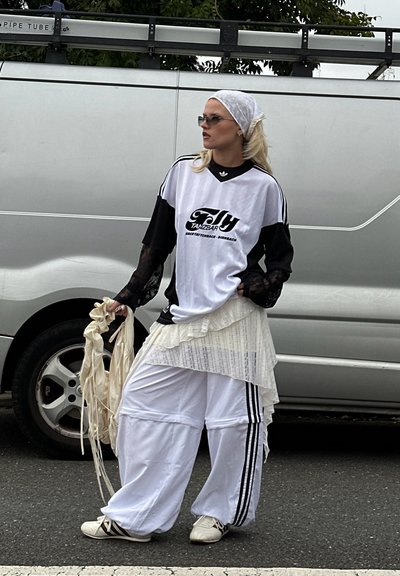 Camisa deportiva negra y blanca de gran tamaño con logo, falda blanca en varias capas, pantalones blancos holgados con rayas negras y zapatos blancos. Los accesorios incluyen un pañuelo en la cabeza y gafas de sol.