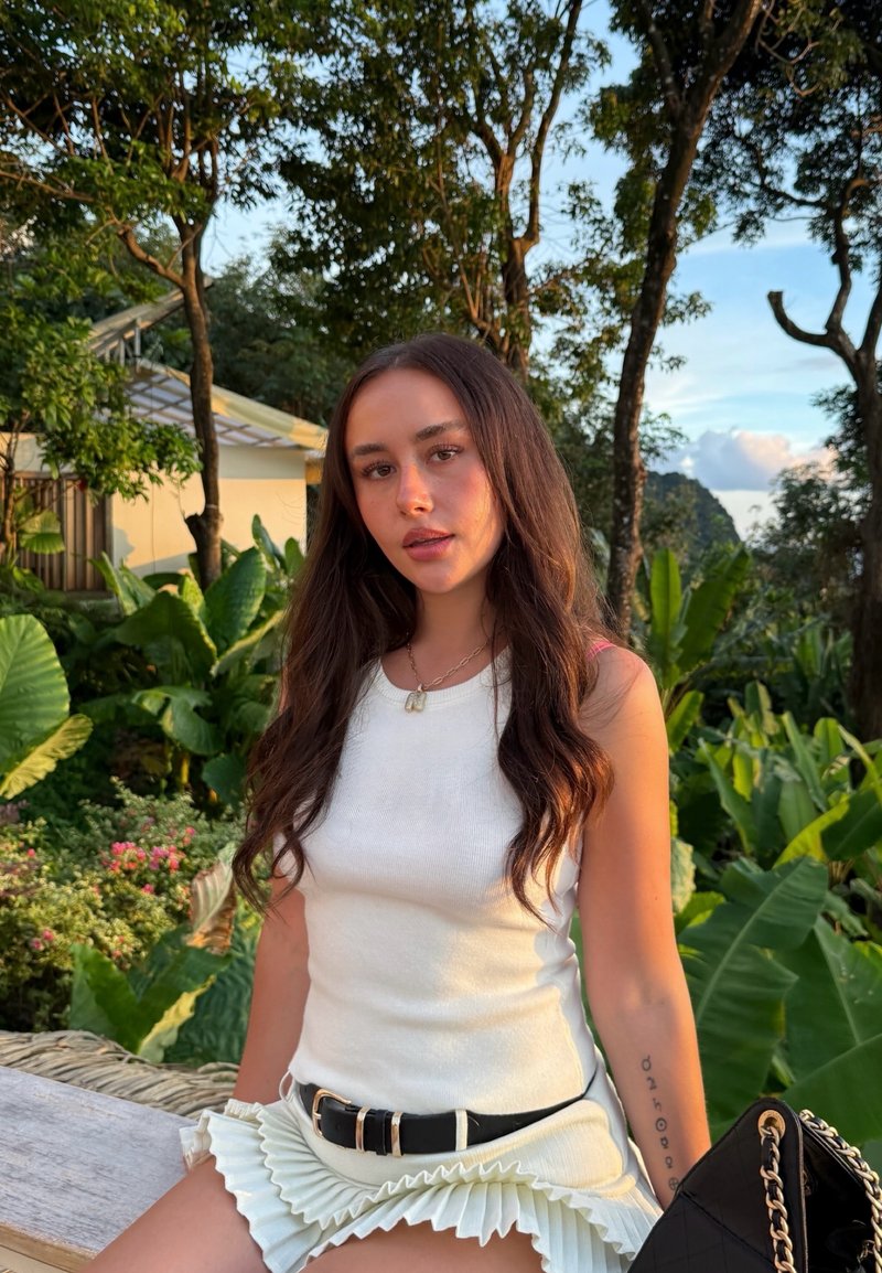 White knit tank top and pleated creamy skirt. Black belt, gold chain necklace. Surrounded by lush green foliage. Natural lighting.