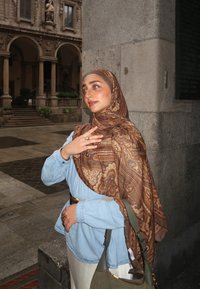 Eine Frau mit einem gemusterten braunen Schal über einem hellblauen Hemd, die eine grüne Tasche hält, mit hellfarbigen Hosen und einem Ring an ihrer Hand.