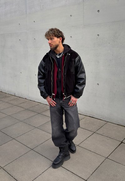 Hombre con cabello rizado vestido con una chaqueta negra y burdeos, botas negras y jeans oscuros y anchos, de pie sobre un suelo de baldosas junto a una pared de hormigón.