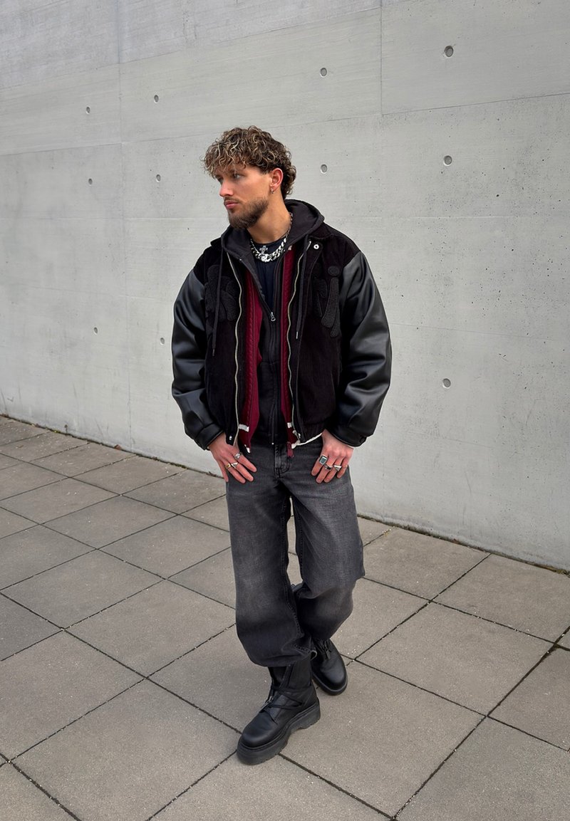 Homme aux cheveux bouclés portant une veste noire et bordeaux, des bottes noires et un jean large foncé, debout sur un sol carrelé près d'un mur en béton.