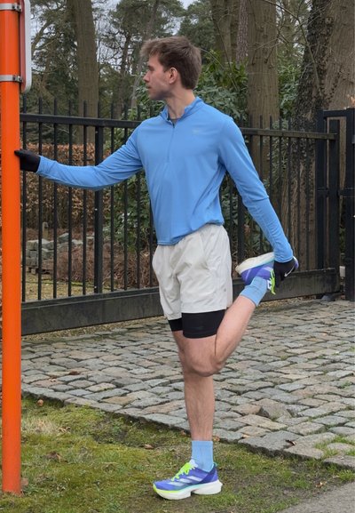 Vestido con una camiseta de manga larga azul claro, pantalones cortos blancos con detalles en negro y zapatillas de correr azul-verdosas, una persona se estira contra un poste.
