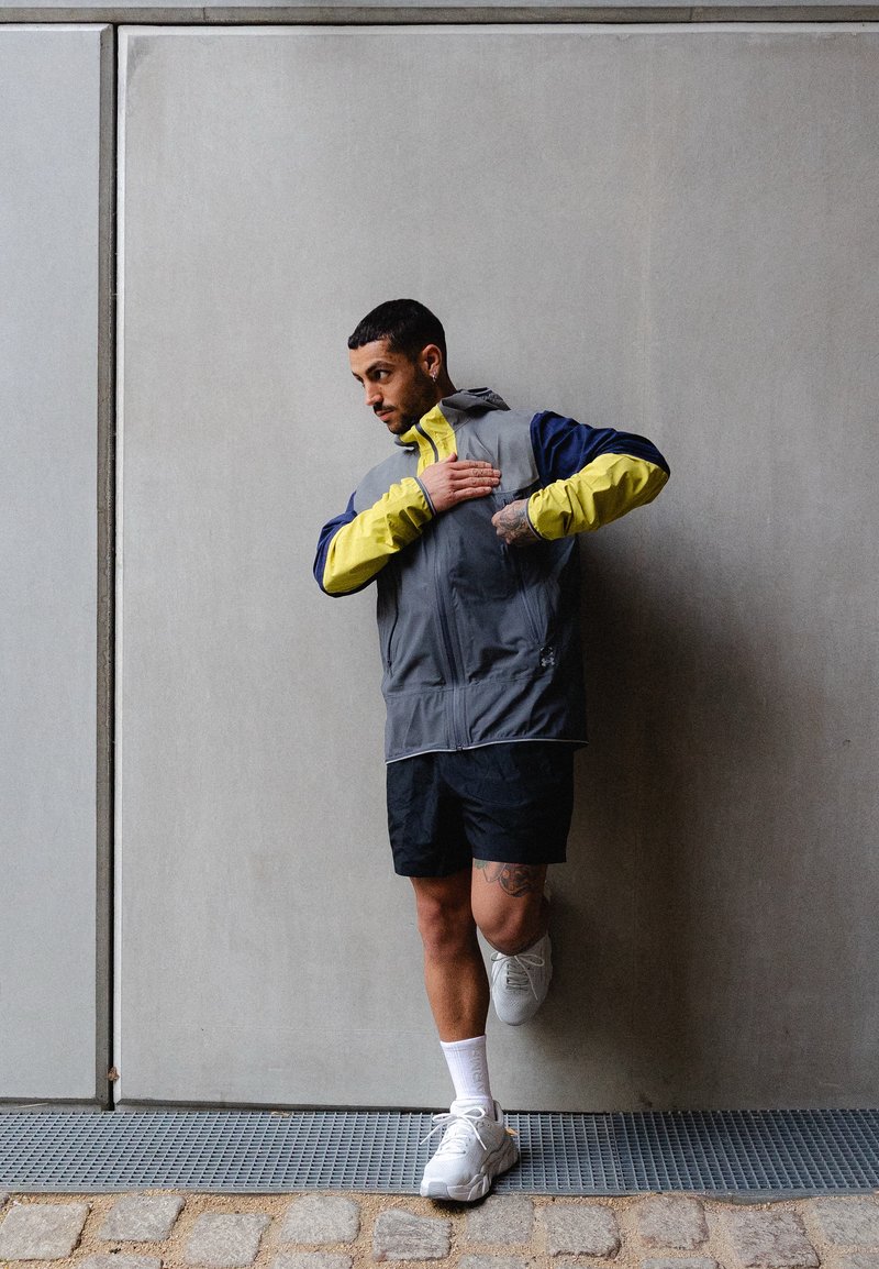 Mann in grauer und gelber Jacke und schwarzen Shorts dehnt sein Bein gegen eine Betonwand im Freien.