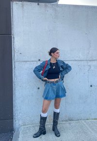 Jeans jakna in krilo z gubami, črn crop top, sivi škornji do kolen, rdeča torba, srebrni dodatki proti betonski zadaj.