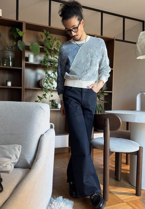 Mann mit Brille und lockigem Haar, der einen gemusterten Pullover und schwarze Hosen trägt, steht neben einem Stuhl in einem modernen Wohnzimmer.