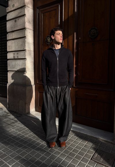 Hombre de pie al sol junto a una puerta de madera, vestido con una chaqueta negra con cremallera, pantalones anchos oscuros y zapatos marrones, manos en los bolsillos.