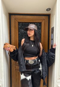 Sort dunjakke, sort cropped top, bukser med ko-mønster. Brun kasket, brun håndtaske med gyldent metalspænde, sølv bæltespænde, langt sort hår.