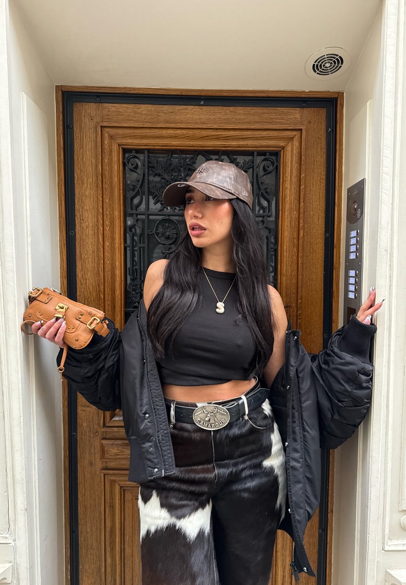 Veste matelassée noire, crop top noir, pantalon à motif vache. Casquette marron, sac à main marron avec ferrures dorées, boucle de ceinture argentée, longs cheveux noirs.