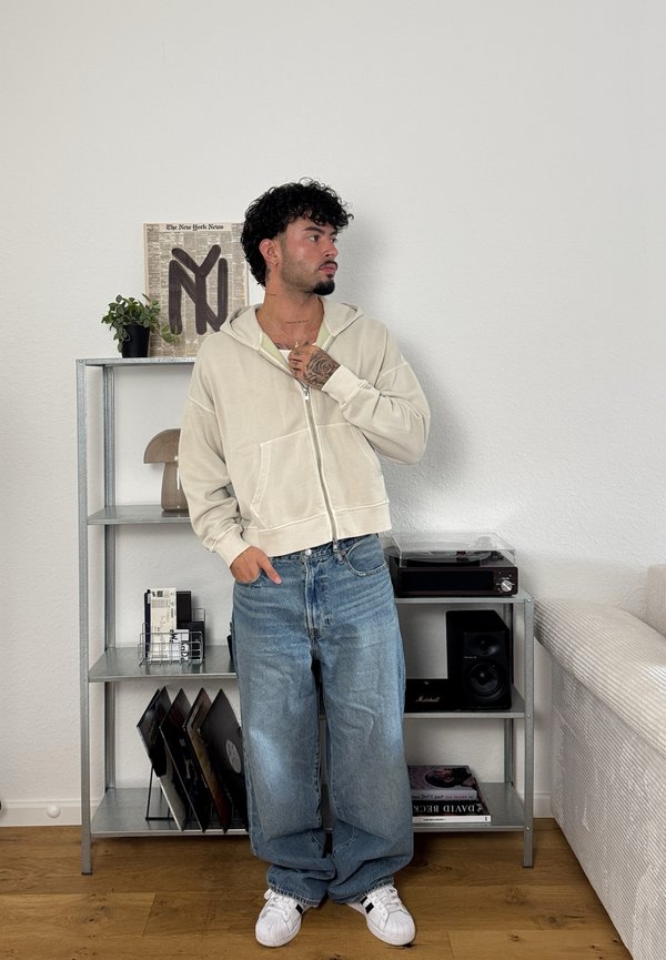 Felpa crema con zip e vestibilità rilassata, abbinata a jeans baggy blu chiaro. Indossata con sneakers bianche, presenta uno sfondo testurizzato con uno scaffale.