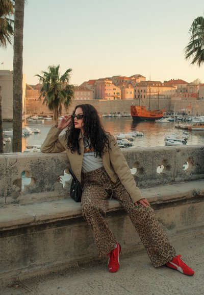 Chaqueta beige, camiseta gráfica blanca, pantalones de leopardo y zapatillas rojas. Los accesorios incluyen gafas de sol oscuras y un bolso negro pequeño. Entorno urbano junto al agua.