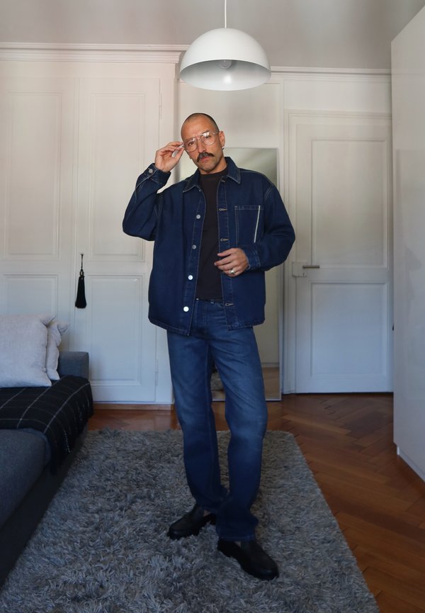 Man with glasses and mustache wearing dark denim jacket and jeans stands on a gray rug in a modern living room with white walls.