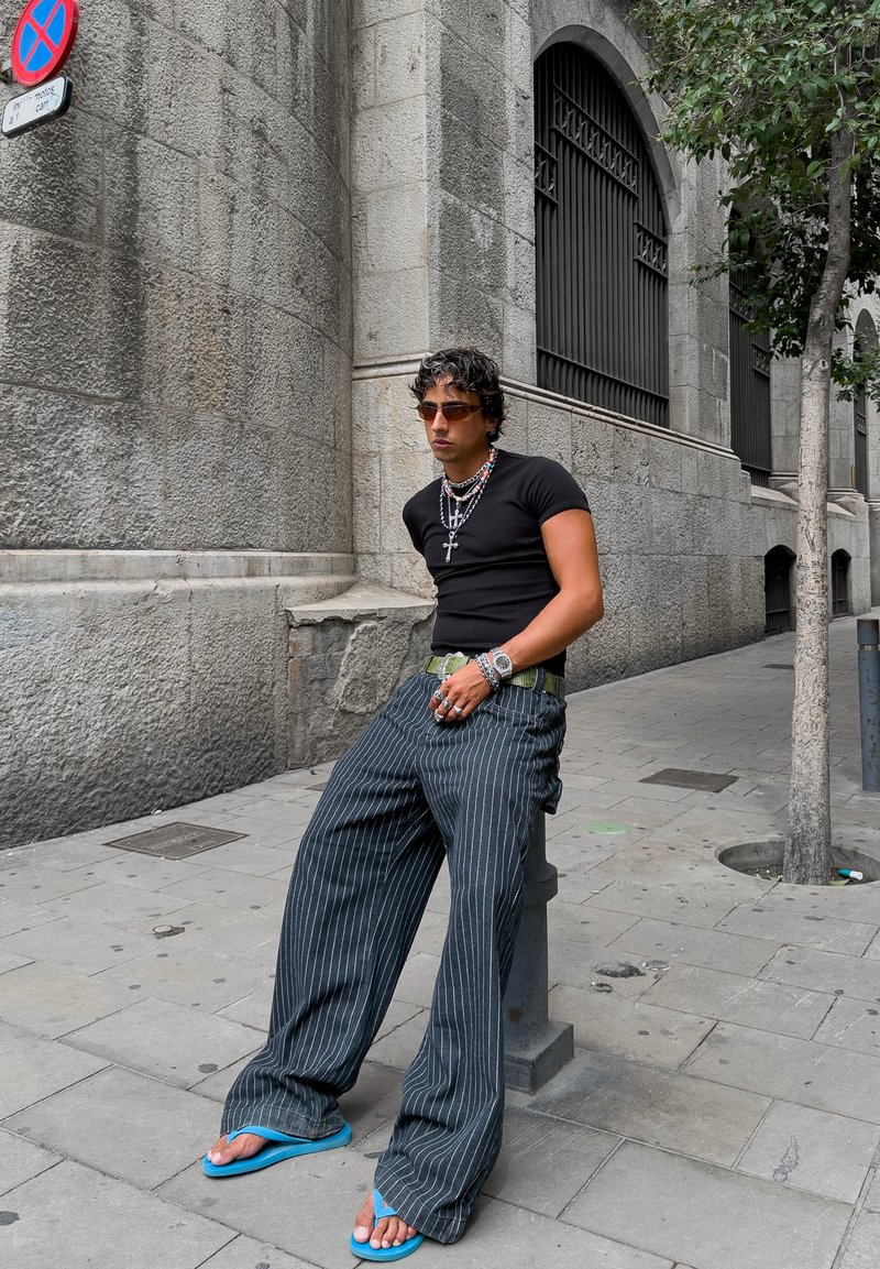 T-shirt noir ajusté, pantalon large à rayures, tongs bleues, colliers superposés, lunettes de soleil, bagues en argent, fond urbain avec mur en pierre.