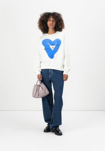 Sudadera blanca con gráfico de corazón azul y texto negro, combinada con jeans de denim azul sueltos y un pequeño bolso de lavanda. Zapatos negros completan el look.