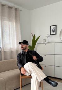 Muž s okuliarmi, čiernou čiapkou, čiernou koženou bundou a bielymi nohavicami, sediaci skrížený na drevenej stoličke v minimalistickej obývacej izbe.