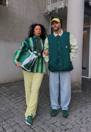 Dos personas están de pie sobre adoquines. Una lleva una camisa verde con un patrón, una corbata verde y pantalones amarillos de textura; la otra viste un chaleco acolchado verde sobre una camisa clara.