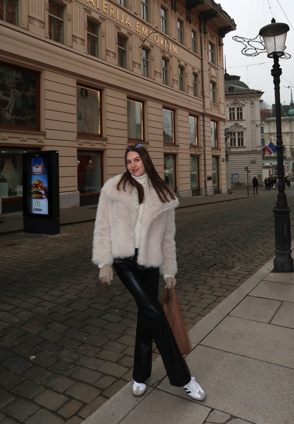 Veste en fausse fourrure rose clair, pull épais blanc, pantalon en cuir noir, et baskets blanches. Sac en papier marron à la main, rue pavée.
