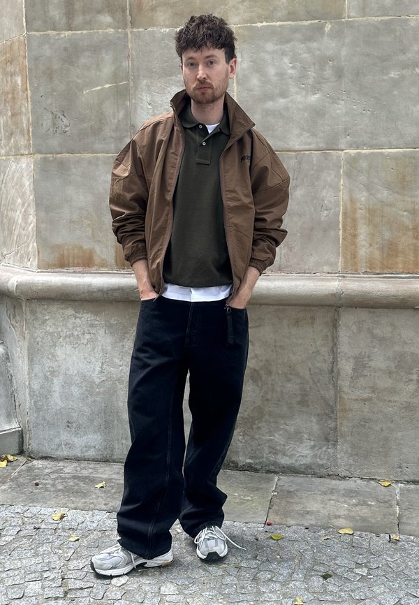 Homme aux cheveux bouclés foncés se tient contre un mur de pierre, portant une veste marron, un polo vert foncé, un pantalon large noir et des baskets grises, les mains dans les poches.