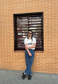 Cămașă polo albă cu accente negre și roșii, blugi din denim albastru și pantofi negri lucioși. Stând în fața unui zid de cărămidă cu o fereastră.