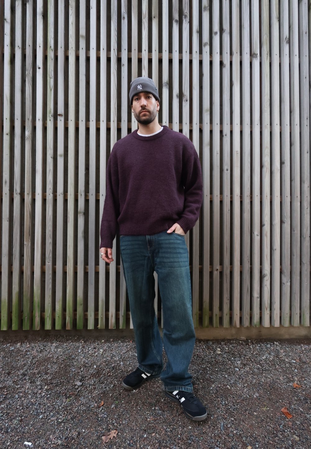 Pull bordeaux à col rond, jean bleu clair et chaussures noires avec des accents blancs, sur fond de lattes en bois.