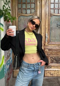 Sort blazer, neon gul kort top med pink tekst, lys blå høj-tskæring jeans og solbriller; holder en kaffekop. Kæde accenter synlige.