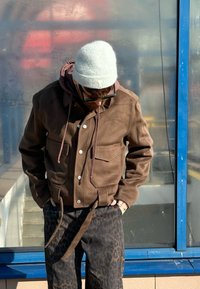 Brown suede jacket featuring two front pockets, a drawstring waist, and white buttons. Layered over a maroon hoodie and paired with black leopard-print trousers.