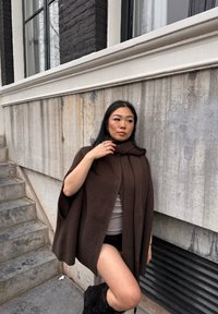 Cape en tricot côtelé marron avec un col haut, dotée de deux poches latérales. La personne qui la porte est assortie d'un haut clair et de shorts sombres. Fond urbain.