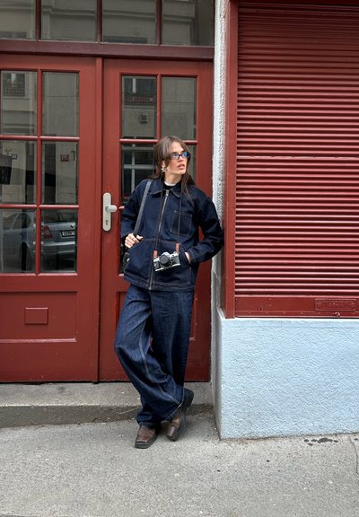 Chaqueta de mezclilla oscura con pantalones de pierna ancha a juego, ambos con costuras en contraste. Los accesorios incluyen una cámara y gafas con tintado azul.