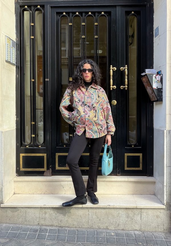 Femme aux cheveux bouclés portant une veste à motifs et un pantalon noir, debout sur des marches devant une porte noire, tenant un sac à main bleu.