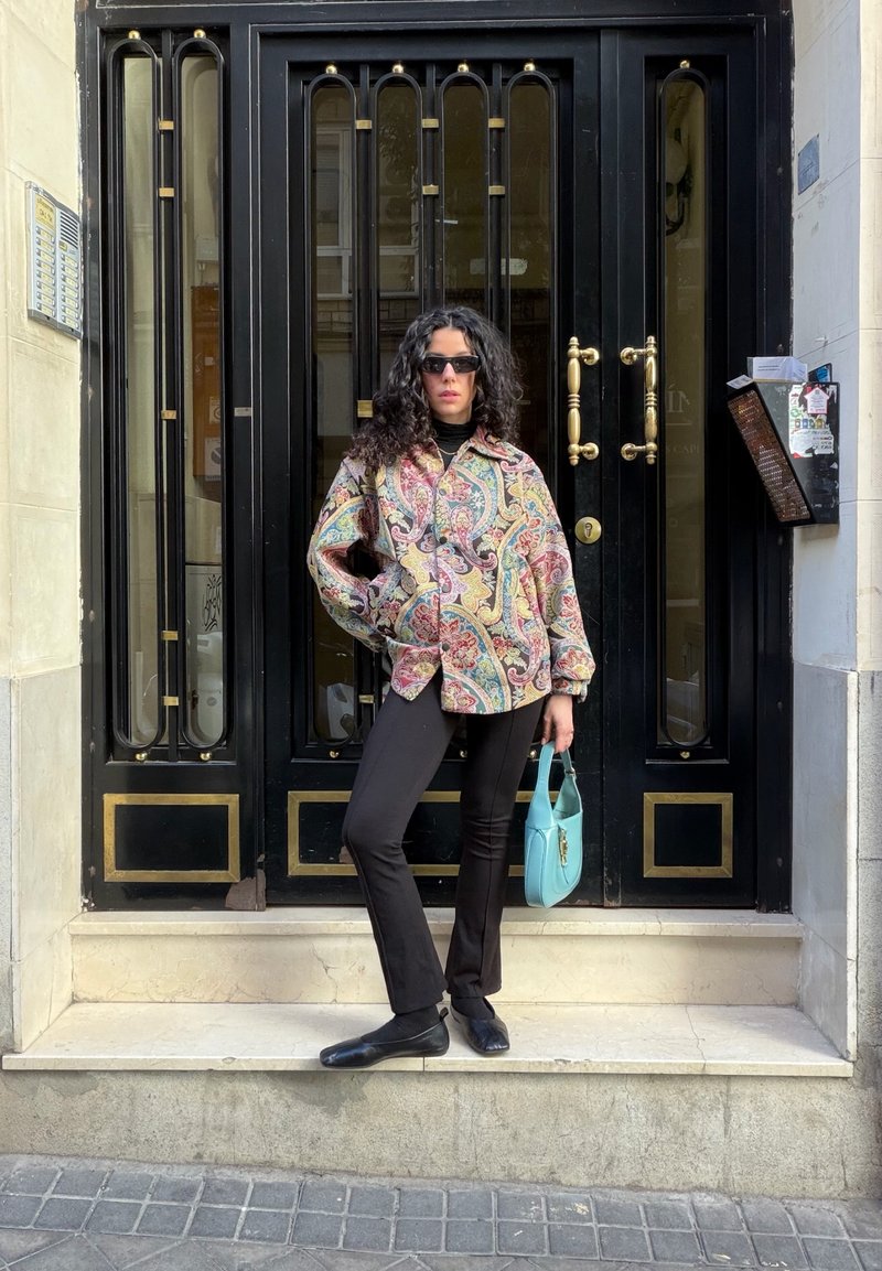 Donna con capelli ricci che indossa una giacca a fantasia e pantaloni neri, in piedi su dei gradini davanti a una porta nera, che tiene in mano una borsa blu.
