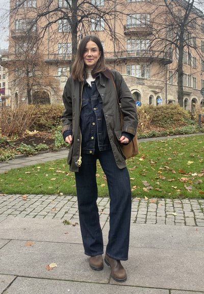 Chaqueta impermeable verde oscuro, peto de mezclilla azul, pantalones anchos azul oscuro, botines marrones, bolso marrón claro, de pie al aire libre con hojas de otoño.