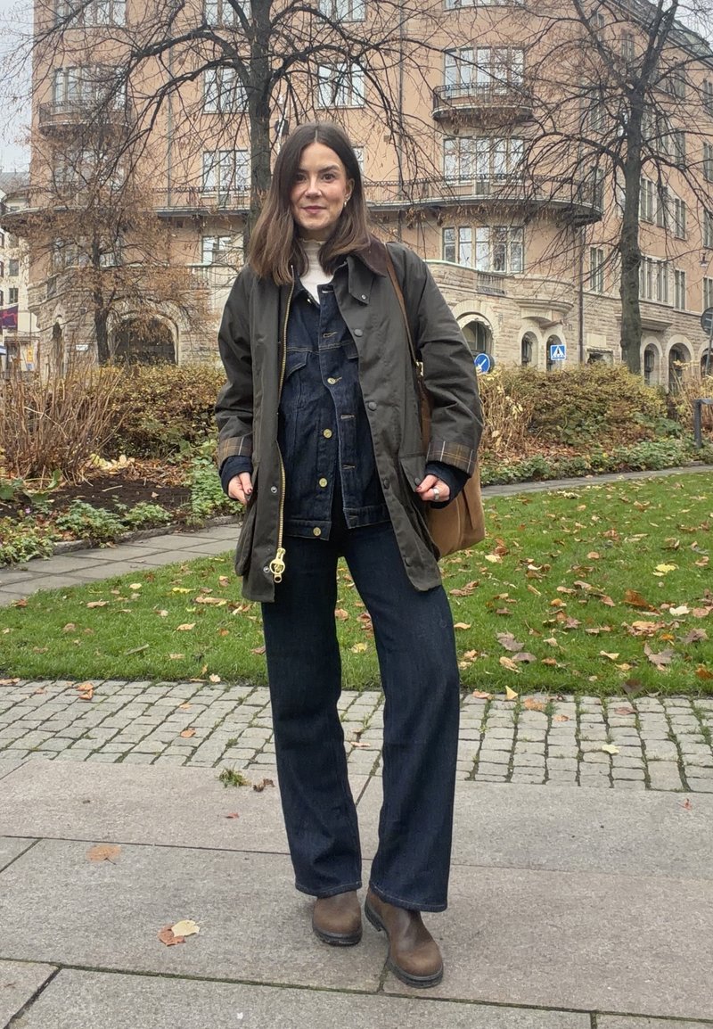 Dunkelgrüne wasserdichte Jacke, blaue Jeanslatzhose, dunkelblaue Hose mit weitem Bein, braune Stiefeletten, hellbraune Tasche, stehend im Freien mit Herbstlaub.