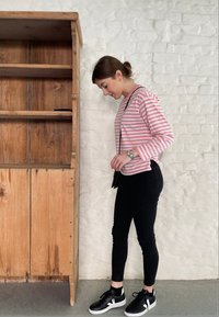 Triibulise, roosa ja valge pikaaelise särgi all on mustad kitsad teksad ja mustad ketsid valgete detailidega, mis asuvad puidust riiuli kõrval.