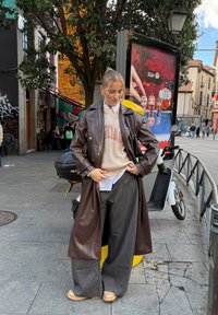 Mulher com casaco comprido de couro castanho, camisola bege, calças largas às riscas finas e sapatos bege, em pé perto de uma scooter e de um cartaz da série Stranger Things numa rua da cidade.