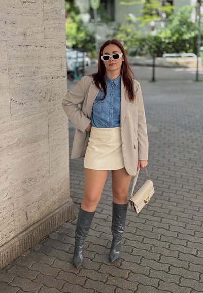 Blazer beige sobre una camisa de mezclilla, falda corta blanca, botas altas grises y gafas de sol blancas. Bolso beige con herrajes dorados.