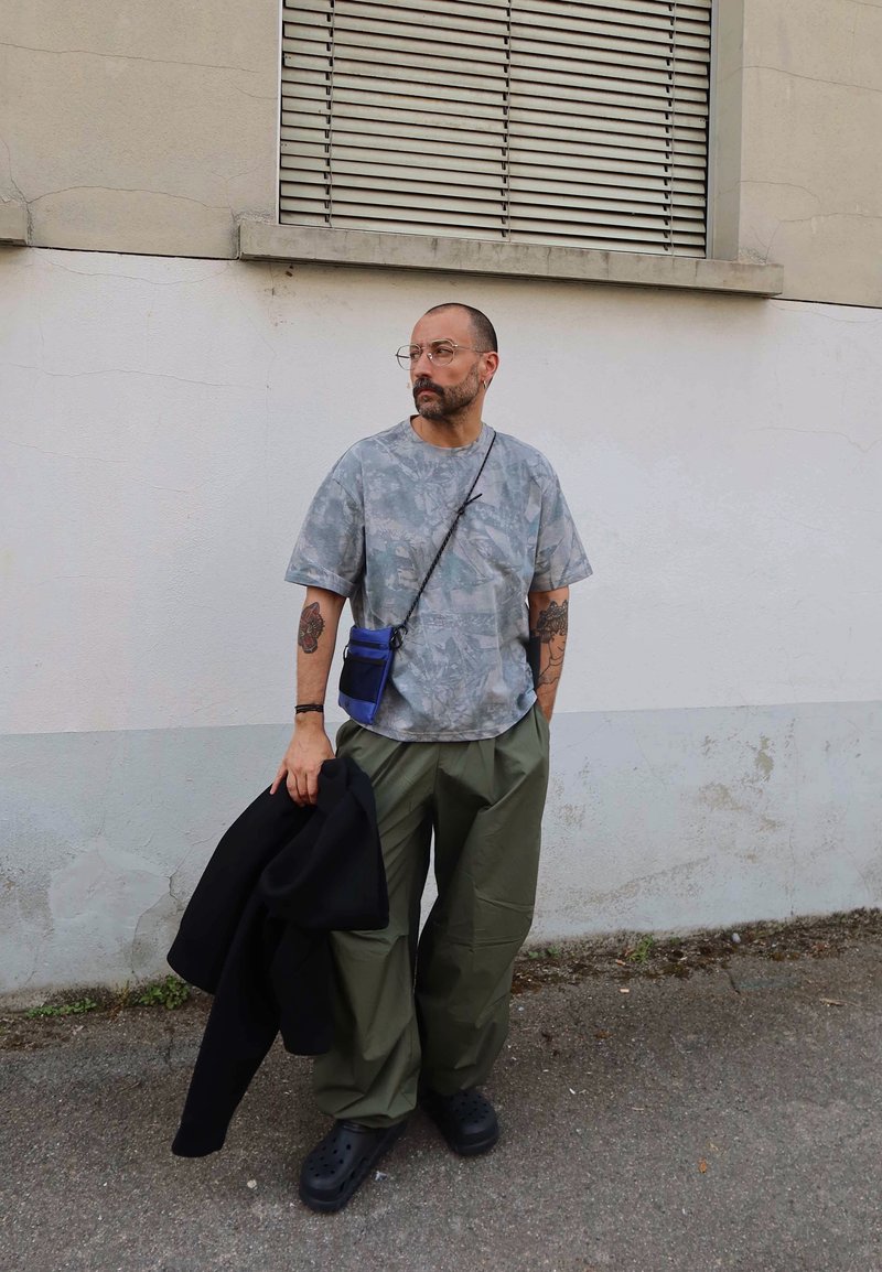 Homme avec des lunettes et des tatouages, portant une chemise grise à motifs et un pantalon olive, tenant une veste noire, debout contre un mur blanc.